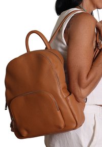 Una donna indossa un top bianco senza maniche e pantaloni, mentre porta uno zaino in pelle marrone tessurizzato di dimensioni medie, dotato di tasche frontali e laterali.