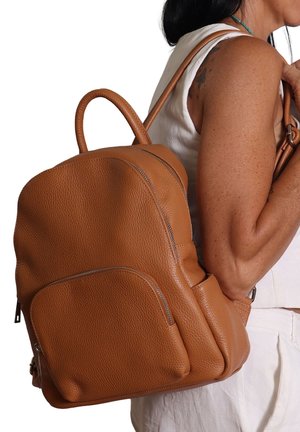 Femme portant un top sans manches blanc et un pantalon, portant un sac à dos en cuir brun texturé de taille moyenne avec des poches avant et latérales.