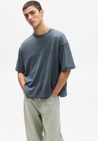 Modré tričko s kulatým výstřihem vyrobené z bavlny s krátkými rukávy, v kombinaci s volnými světle zelenými džíny. Minimalistický design, bez viditelných vzorů.