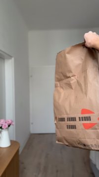 Ruda popierinė pakuotė, laikoma aukštyn, su ryškia oranžine grafika ir horizontaliai atspausdintais brūkšniniais kodais šone. Matyti raukšlėta tekstūra.