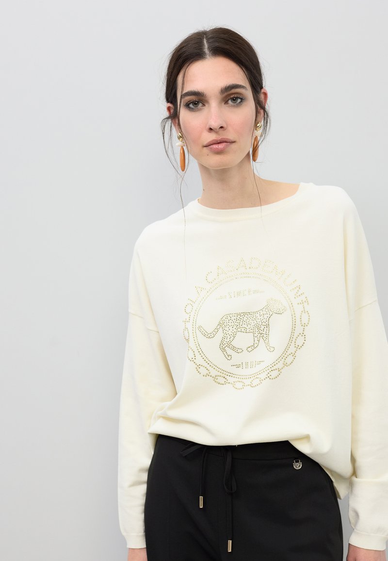 Sweat-shirt blanc à col rond et épaules tombantes, avec un imprimé léopard aux accents dorés. Associé à un pantalon noir.