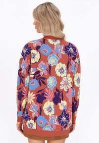 Donna con capelli biondi indossa un maglione color ruggine con grandi motivi floreali blu, gialli, rosa e viola, fotografata di spalle.