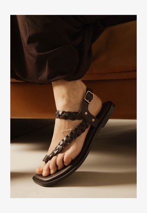 Pie que lleva una sandalia de cuero marrón oscuro trenzada con hebilla ajustable, apoyado cerca de un sofá marrón sobre un suelo claro.