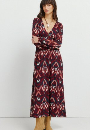 Vrouw met donker haar die een lange mouw, maxi-jurk met een patroon in rood, marineblauw en wit draagt, gecombineerd met suède laarzen in een tan kleur en meerdere kettingen.