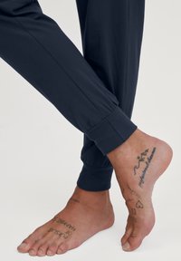 Marine joggerbroeken met elastische manchetten, met een blote voet waarop zichtbare tatoeages aan de bovenkant en zijkant van de voet te zien zijn. Zachte, gladde stofstructuur.