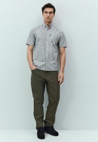 Camicia a maniche corte con bottoni, motivo a quadretti blu e bianchi, abbinata a pantaloni verde oliva e scarpe nere in camoscio.