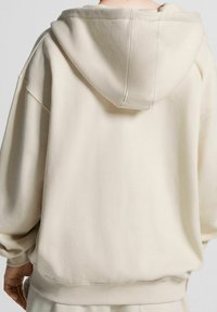 Persona che indossa una felpa beige oversize vista di spalle, con il cappuccio abbassato e le mani rilassate lungo i fianchi.