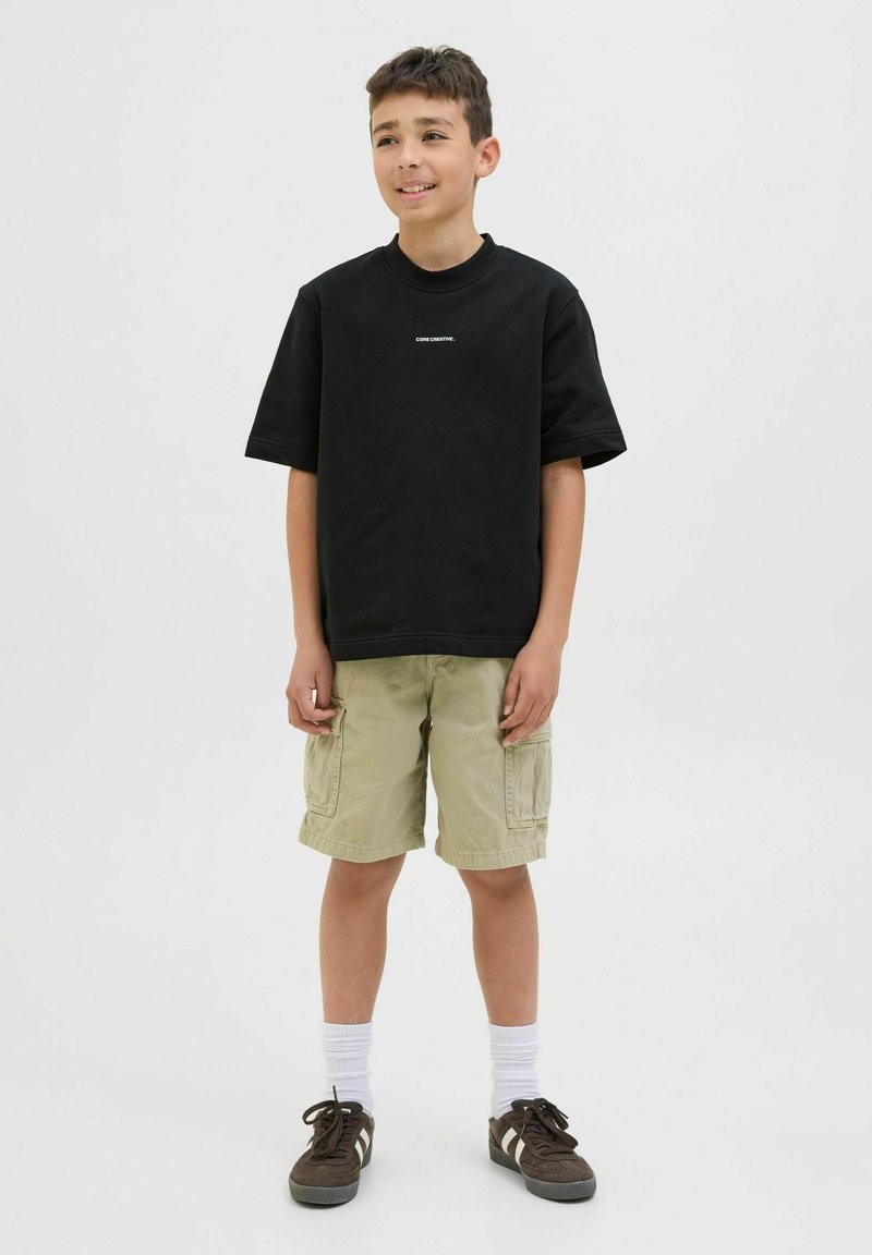 Maglietta nera a maniche corte con testo bianco, abbinata a pantaloni cargo beige. Sneakers marroni e calzini bianchi completano l'outfit. Sfondo neutro.