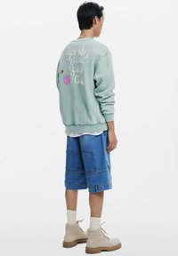 Sweatshirt vert menthe avec texte et motifs brodés dans le dos, associé à un short en jean bleu et des bottes beiges à semelle épaisse.