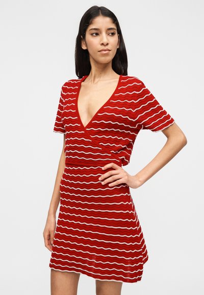 Frau trägt ein rot-weiß gestreiftes, kurzärmeliges Kleid mit wellenförmigen horizontalen Streifen, posiert mit einer Hand in der Hüfte und schaut zur Seite.