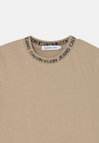 T-shirt beige à manches courtes avec un col rond et un logo noir "Calvin Klein Jeans" sur le col. Fabriqué en tissu coton doux.