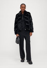 Μαύρο σακάκι από τεχνητό γούνα με διαγώνιες ρίγες, ψηλό γιακά και ασημί φερμουάρ. Φοριέται πάνω από μαύρη μπλούζα και παντελόνι, συνδυασμένο με μια μικρή μαύρη τσάντα.