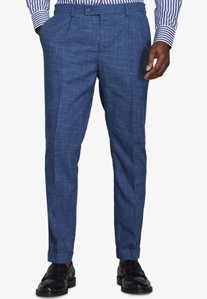 Uomo che indossa pantaloni eleganti blu dal taglio aderente con un motivo a trama incrociata sottile, scarpe eleganti nere e una camicia a righe blu e bianche.