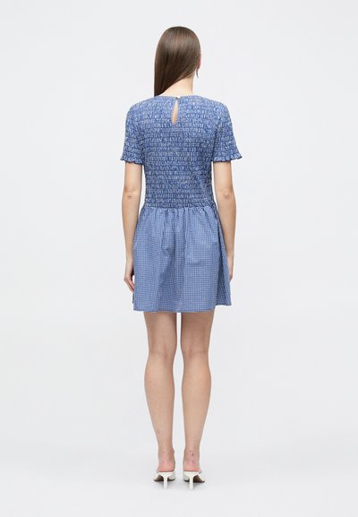 Femme portant une robe à motifs mélangés bleus à manches courtes et des mules blanches à talons, vue de dos sur un fond blanc uni.