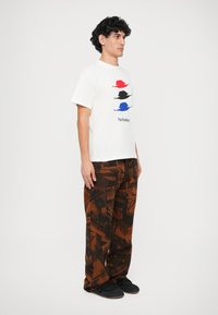 Weißes T-Shirt mit bunten Hutgrafiken in Rot, Schwarz und Blau; kombiniert mit braunen gemusterten Hosen mit dunklen Druckakzenten.