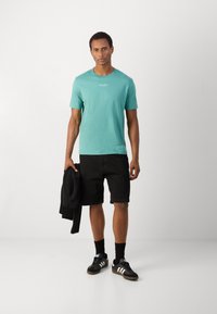 Turquoise színű pamut póló rövid ujjakkal és kerek nyakkivágással, fekete sorttal párosítva. A modell egy fekete dzsekit tart, és fekete sportcipőt visel.