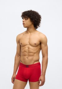 Boxers de algodão vermelhos com uma cintura castanha exibindo o logo "Calvin Klein". O design é ajustado e possui uma textura suave.