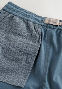 Pantalones cortos azules con un bolsillo de tela estampada con un diseño geométrico, cinturilla elástica y textura suave. La etiqueta dice "NEXT DURABLE GOODS."