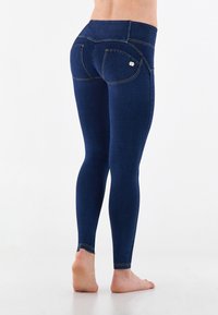 Leggings de mezclilla azul oscuro con una cintura alta, silueta ajustada y costuras amarillas en contraste. Bolsillos traseros con un sutil detalle de marca.