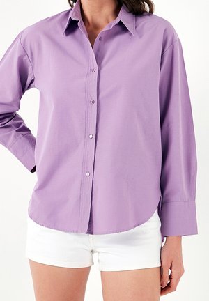 Femme portant une chemise lavande à manches longues boutonnée avec col et un short blanc, debout avec une main sur la hanche.