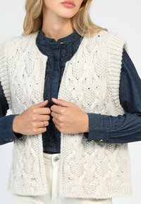 Gilet tricoté blanc chunky avec un motif en cable, à ouverture sur le devant, et des bordures en côtes, porté par-dessus une blouse en denim bleu foncé avec des détails en volants.