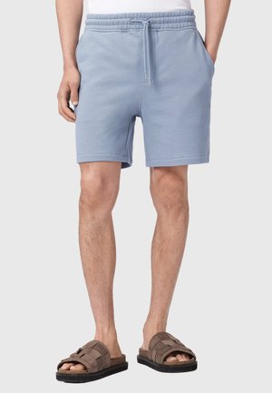Mand iført lyseblå shorts med snøre, forlommer og brune sandaler med åbne tæer mod en ensfarvet baggrund.