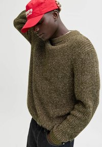 Tekstūruotas alyvuogių žalios spalvos megztinis su plačia iškirpte ir stambiu mezgimo raštu. Dėvimas su raudona kepure, papuošta baltais siuvinėtus tekstais.