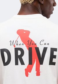 Άνδρας που φοράει λευκό πουκάμισο με μεγάλο κόκκινο περίγραμμα γκόλφερ που κάνει κούνημα και κείμενο "When You Can DRIVE" στην πλάτη.