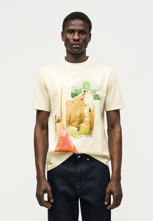 Mand iført en lysebeige t-shirt med farverig grafik af gamle egyptiske statuer og strukturer, kombineret med mørkeblå jeans.