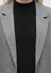 Femme portant un pull à col roulé noir et un blazer gris à motif chevron.