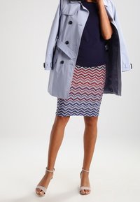 Jasnoszara trench coat z czarnymi guzikami, w połączeniu z spódnicą w zygzakowy wzór w czerwono-biało-niebieskich kolorach oraz nagimi sandałami na obcasie.