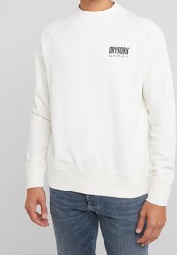 Sudadera blanca con cuello redondo y puños acanalados, que presenta un pequeño logotipo negro en el lado izquierdo del pecho. Textura suave, corte relajado y material elástico.