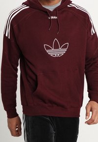 Man som bär en vinröd Adidas-hoodie med vit trefoil-logotyp och tre vita ränder på axlarna, tillsammans med svarta Adidas-byxor.
