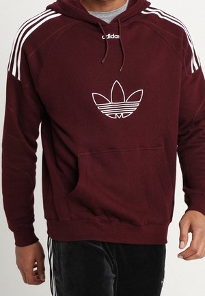 Hombre vestido con sudadera marón de Adidas con logo trefoil blanco y tres franjas blancas en los hombros, combinada con pantalones negros de Adidas.