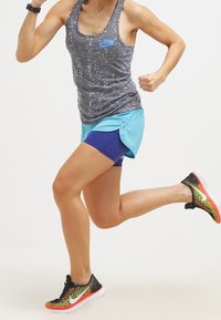 Top sin mangas gris con el logo azul "RUN", pantalones cortos de running en color aqua y zapatillas texturizadas de colores negro y amarillo, en una pose de carrera.