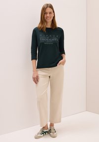 Dunkelgrünes Langarmoberteil mit bedrucktem Text, kombiniert mit hellbeigen weit geschnittenen Hosen und grauen Sneakers mit grünen Akzenten.