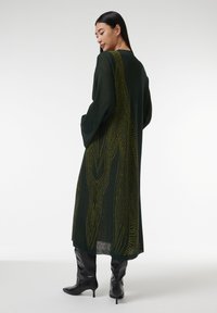 Lang groene jurk met een lichte, doorschijnende textuur, voorzien van opvallende gele lijnpatronen en een bescheiden, relaxed fit. Combineert mooi met glanzende zwarte laarzen.