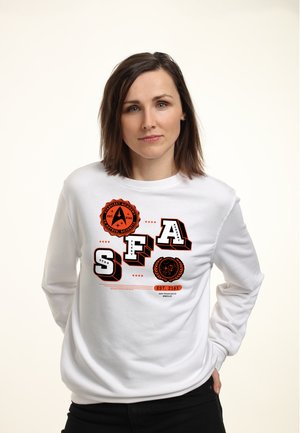 Kobieta w białej bluzie z pomarańczowymi logo Akademii Starfleet i czarnych spodniach, stojąca z rękami w kieszeniach, neutralne tło.