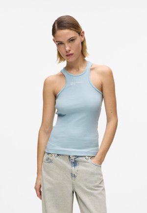 Žena s krátkymi blond vlasmi oblečená v svetlomodrom topánom tanktopu s halter krkom a svetlomodrých džínsoch s vysokým pásom, stojaca s jednou rukou v vrecku.