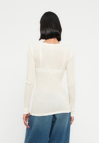 Φωτεινό μπεζ τοπ με μακριά μανίκια σε ριμπ υφή με εφαρμοστή σιλουέτα και λαιμόκοψη με στρογγυλή λαιμόκοψη, συνδυασμένο με τζιν παντελόνι φαρδιάς γραμμής μπλε χρώματος.