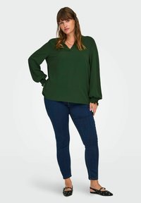 Donna in piedi con una mano sui fianchi, che indossa una blusa a maniche lunghe verde scuro, jeans skinny blu scuro e ballerine nere con occhielli.