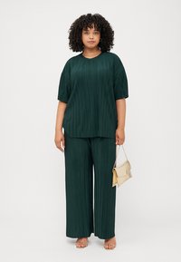 Γυναίκα με σγουρά μαλλιά φοράει ένα πτυχωτό σκούρο πράσινο τοπ και αντίστοιχα παντελόνια με πλατιά πόδια, κρατώντας μια μικρή χρυσή τσάντα, σε έναν απλό φόντο.