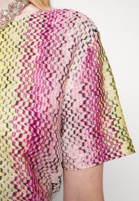 Kortærmet strikket top med et farverigt zigzag-mønster i pink, gul og sort, båret på en persons skulder og overarm.