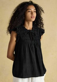 Musthave mustrinna ilma varrukatega pluus, kogutud detailide, volangidega õlavarre servade, lukktagumisega ning lipsuga, valmistatud tekstuurilisest kangast.