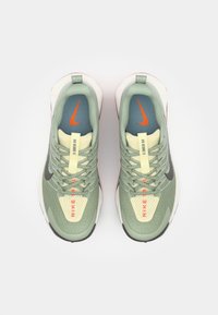 Zeleno-beige tenisky Nike s textúrovanou sieťovanou hornou časťou, oranžovými akcentmi a čiernym logom Swoosh. Majú zaoblenú špičku a mäkkú podrážku.