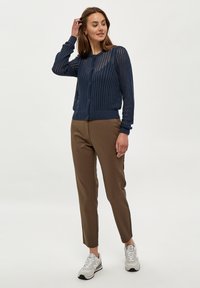 Mornarji pleteni kardigan z teksturiranim odprtim vzorcem, z gumbom na zapiranju, usklajen z rjavimi krojenimi hlačami in sivimi športnimi čevlji.