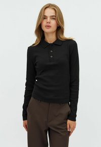Svart långärmad polo tröja med ribbad textur, treknapprad och klassisk krage, ihop med bruna byxor.