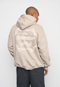Felpa beige con un grande motivo grafico sulla schiena, che presenta vari testi e simboli bianchi. Vestibilità morbida e rilassata con cappuccio.