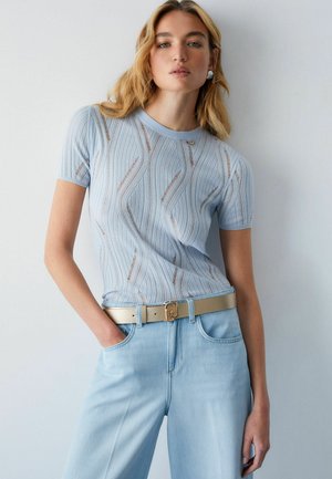 Ženska, oblečena v svetlo modro kratko majico z rebrastim vzorcem in valovitimi linijami, svetlo modre kavbojke, bež pas in bele uhane, stoji ob belem zidu.