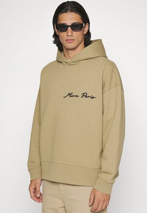 Jonge man met zwarte zonnebril en een beige hoodie met de tekst "Mira Paris", staand tegen een effen lichte achtergrond.